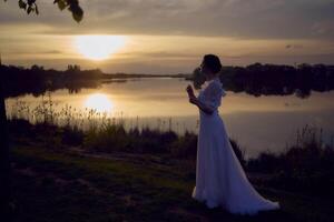 uma mulher dentro uma branco vintage vestir em a costa do uma lago às pôr do sol foto