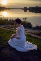 uma mulher dentro uma branco vintage vestir em a costa do uma lago às pôr do sol foto