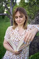 mulher dentro uma pastel vestir lê uma livro perto uma árvore dentro a jardim foto