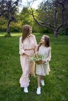 uma mãe e filha dentro pastel vestidos andar através a jardim com uma cesta do flores foto