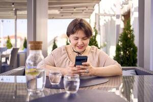 mais Tamanho mulher dentro pêssego penugem vestir escolhe uma prato dentro uma restaurante através da qr código dentro Smartphone, água com limão e óculos dentro frente do dela foto