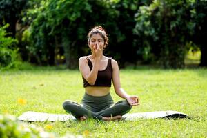 elegante mulher sentado Cruz de pernas e fazendo respiração exercício dentro ensolarado parque foto