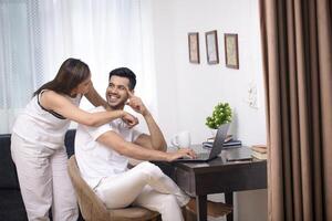 cenário do indiano casal sorridente e marido sentado em cadeira e esposa em pé ambos estão olhando às cada de outros foto