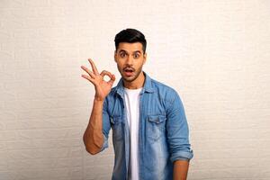 indiano masculino modelo vestindo jeans Jaqueta e fazer OK placa com dedos foto