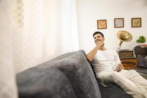 homem dentro branco camiseta sorridente segurando branco caneca dentro mão enquanto sentado em sofá foto