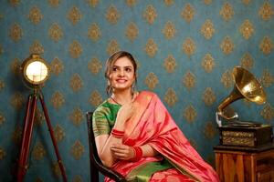 sorridente mulher dentro vermelho saree sentado em uma cadeira e posando dentro frente do a Câmera foto