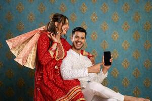 uma casal vestido tradicionalmente rindo dentro frente do a Câmera enquanto segurando compras bolsas e uma celular foto