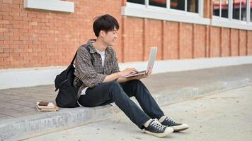 uma feliz ásia masculino Faculdade aluna senta em a rua perto a campus construção usando dele computador. foto