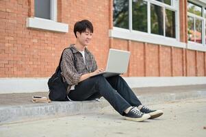 uma feliz ásia masculino Faculdade aluna senta em a rua perto a campus construção usando dele computador. foto