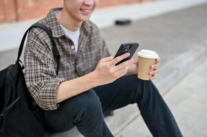 uma jovem masculino Faculdade aluna senta em a rua usando dele Smartphone e segurando uma café copo. foto