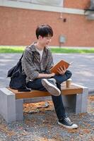 uma feliz, relaxado ásia masculino Faculdade aluna senta em uma Banco dentro a campus parque lendo uma livro didático. foto