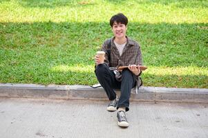 sorridente ásia aluna senta em a rua dentro a campus parque com uma café copo e uma livro dentro dele mão foto