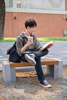 uma feliz ásia masculino Faculdade aluna é desfrutando dele café e lendo uma livro em uma banco. foto