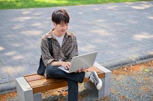 uma jovem ásia masculino Faculdade aluna usando dele computador portátil computador em uma Banco dentro a campus parque. foto