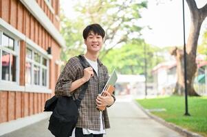 uma alegre ásia homem aluna carrinhos lado de fora uma tijolo construção em campus, segurando uma computador portátil e livros. foto