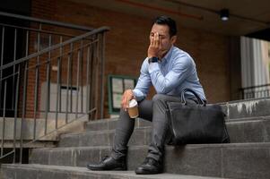 uma estressado ásia homem de negocios, aparecendo cansado e infeliz, senta em a passos lado de fora uma prédio. foto