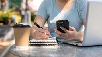 uma mulher olhando às dela Smartphone e levando notas dentro dela caderno às uma mesa ao ar livre. foto