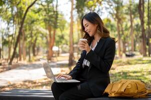 a elegante ásia empresária trabalhando remotamente às uma parque, bebericando café e trabalhando em dela computador portátil. foto
