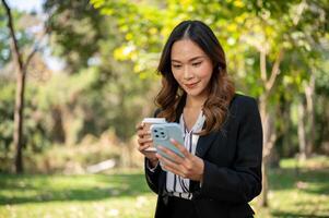 uma empresária é bebericando café e verificação mensagens em dela Smartphone enquanto relaxante dentro uma parque. foto