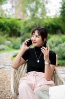 uma positivo ásia mulher relaxante dentro uma verde jardim, sentado às uma mesa e falando em a telefone. foto