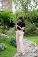 uma positivo ásia mulher usando dela telefone, conversando ou verificação mensagens enquanto caminhando dentro uma jardim. foto