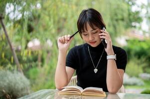 a atraente ásia mulher é levando notas dentro uma livro enquanto falando em a telefone dentro uma verde jardim. foto
