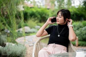 uma positivo ásia mulher relaxante dentro uma verde jardim, sentado às uma mesa e falando em a telefone. foto