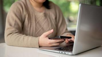 uma mulher segurando uma Smartphone enquanto digitando em dela computador portátil teclado, trabalhando em dela computador às uma mesa foto