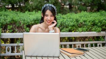 a atraente, positivo ásia mulher trabalhando em dela computador portátil computador às uma mesa dentro uma verde jardim. foto