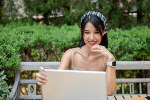 a atraente, positivo ásia mulher trabalhando em dela computador portátil computador às uma mesa dentro uma verde jardim. foto
