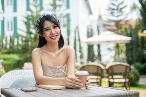 uma lindo ásia mulher senta às uma mesa com uma livro e uma café copo em a mesa dentro uma jardim. foto