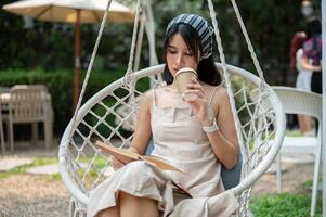 uma mulher é bebericando café e concentrando em lendo uma livro enquanto arrepiante em uma balanço dentro a jardim. foto