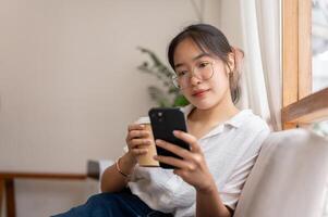 uma mulher lendo conectados blog em dela Smartphone enquanto bebericando café em a sofá dentro a vivo sala. foto