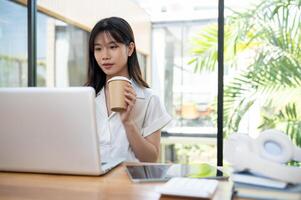 uma lindo ásia empresária bebericando café enquanto concentrando em dela trabalhos em uma computador portátil computador. foto