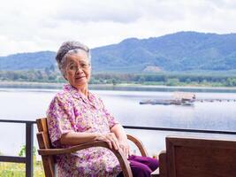 Senior mulher com curto cinzento cabelo, vestindo copos, sorridente e olhando às a Câmera enquanto sentado em uma de madeira cadeira lado a lago. espaço para texto. conceito do envelhecido pessoas e relaxamento foto