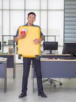 uma jovem homem carregando grande amarelo bagagem, sorridente e olhando às a Câmera enquanto em pé dentro a escritório. preparar para a próximos feriado. feriado conceito foto