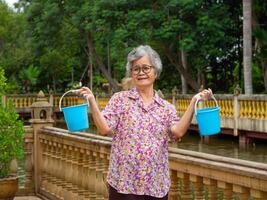 Senior mulher segurando azul plástico baldes, sorridente e olhando às a Câmera enquanto em pé dentro a parque. espaço para texto. conceito do envelhecido pessoas e relaxamento foto