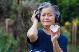 Senior mulher com curto cinzento cabelo, vestindo sem fio fones de ouvido, segurando uma em forma de coração branco cerâmica, e olhando às a Câmera enquanto em pé dentro uma jardim. conceito do envelhecido pessoas e relaxamento foto