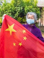 retrato do idosos ásia mulher vestindo uma face mascarar e segurando a China bandeira enquanto em pé dentro uma jardim. mascarar para proteção vírus, COVID-19, coronavírus. conceito do envelhecido pessoas e cuidados de saúde foto