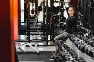 lindo menina com mãos em halteres olhando em dela reflexão dentro a espelho dentro uma Esportes corredor. fino jovem mulher é levando halteres dentro frente do uma espelho dentro ginástica clube foto