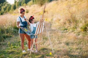 beleza mãe pintura com dela pequeno filha. à moda mulher desenhando a cenário com pequeno garota. fofa criança dentro uma branco camiseta e azul jeans. mãe dia. foto