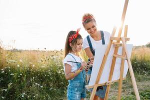 beleza mãe pintura com dela pequeno filha. à moda mulher desenhando a cenário com pequeno garota. fofa criança dentro uma branco camiseta e azul jeans. mãe dia. foto