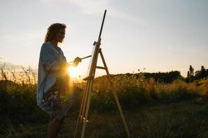 silhueta do uma Loiras menina artista. senhora tintas uma pintura em a tela de pintura com a Socorro do tintas. uma de madeira cavalete mantém a cenário. verão é uma ensolarado dia, pôr do sol. foto