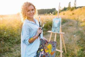bonita mulher é pintura. aberto ar sessão. fofa mulher desenha uma cenário às pôr do sol. menina artista. foto