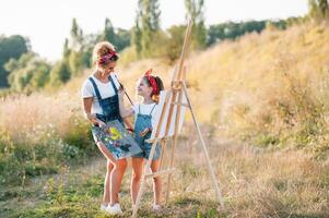 beleza mãe pintura com dela pequeno filha. à moda mulher desenhando a cenário com pequeno garota. fofa criança dentro uma branco camiseta e azul jeans. mãe dia. foto