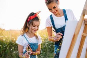 beleza mãe pintura com dela pequeno filha. à moda mulher desenhando a cenário com pequeno garota. fofa criança dentro uma branco camiseta e azul jeans. mãe dia. foto