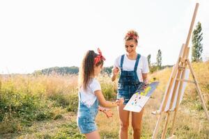 beleza mãe pintura com dela pequeno filha. à moda mulher desenhando a cenário com pequeno garota. fofa criança dentro uma branco camiseta e azul jeans. mãe dia. foto