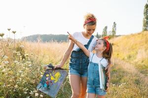 beleza mãe pintura com dela pequeno filha. à moda mulher desenhando a cenário com pequeno garota. fofa criança dentro uma branco camiseta e azul jeans. mãe dia. foto