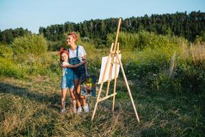 beleza mãe pintura com dela pequeno filha. à moda mulher desenhando a cenário com pequeno garota. fofa criança dentro uma branco camiseta e azul jeans. mãe dia. foto