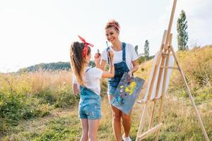 beleza mãe pintura com dela pequeno filha. à moda mulher desenhando a cenário com pequeno garota. fofa criança dentro uma branco camiseta e azul jeans. mãe dia foto